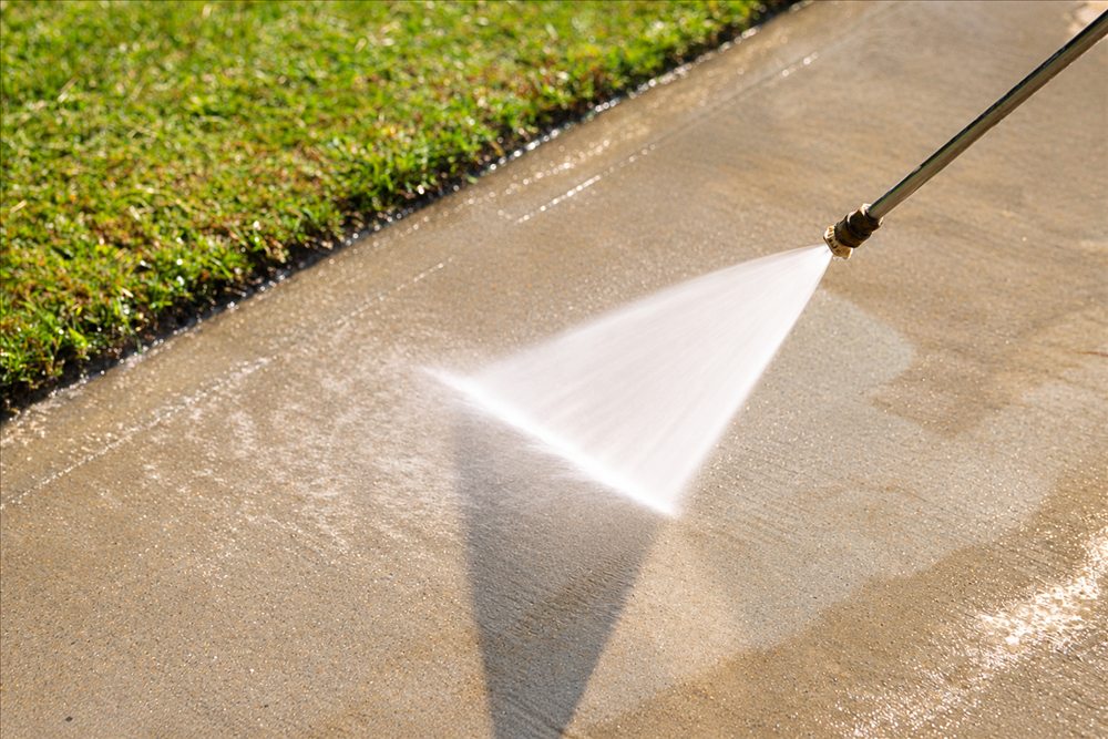 Pressure washer wand spraying a concrete path beside a lawn